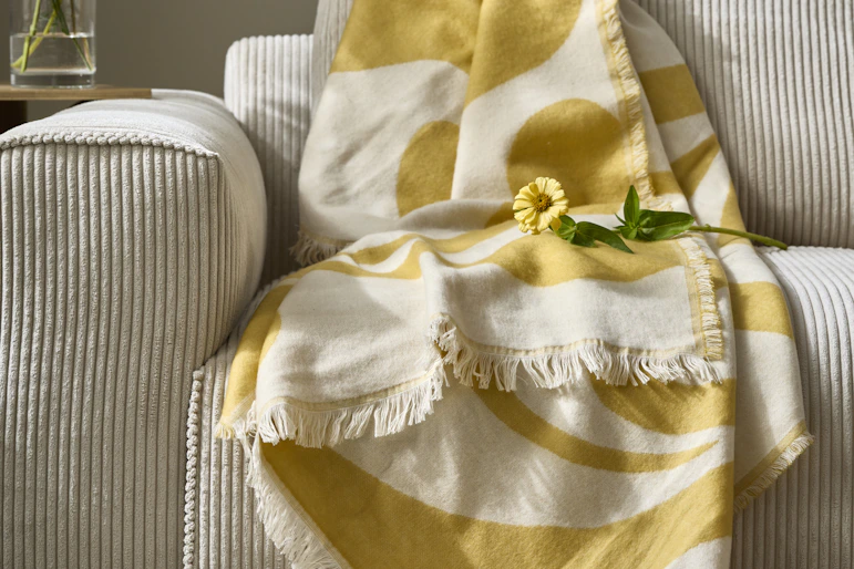 I en beige soffa ligger en tunn, gul filt. Ovanpå filten ligger en gul blomma