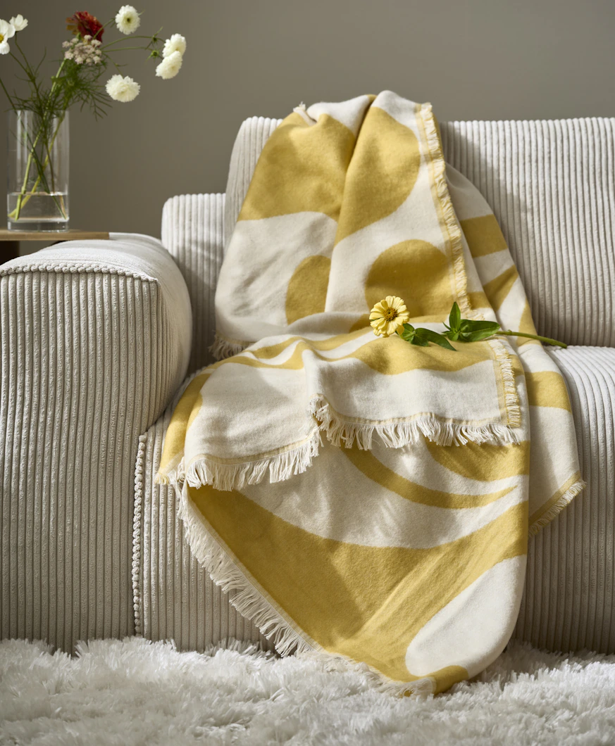 I en beige soffa ligger en tunn, gul filt. Ovanpå filten ligger en gul blomma