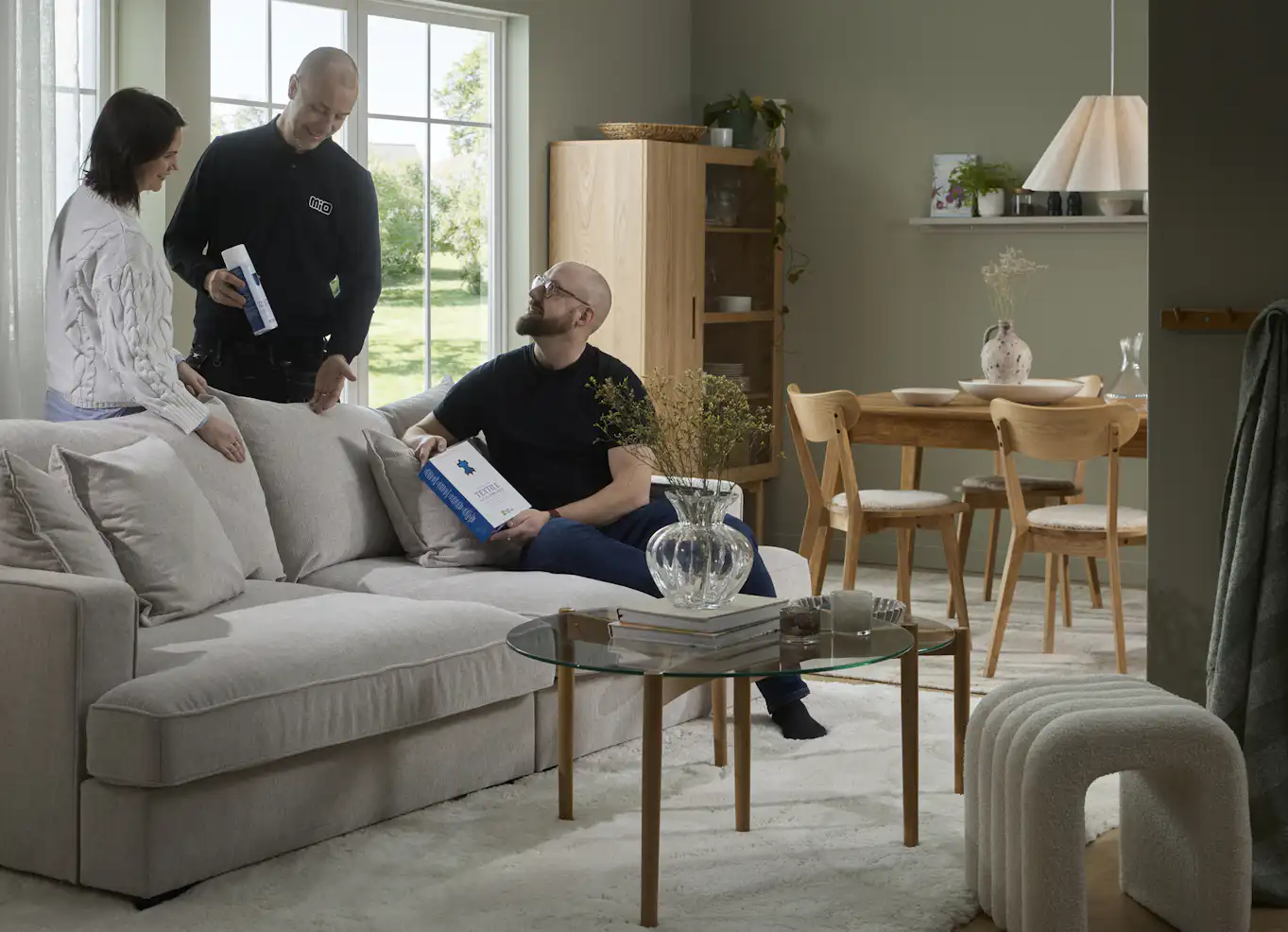 Servicepersonal från Mio med svarta kläder står i vardagsrum och pratar om möbelvårdsprodukter med kvinna intill och en man som sitter i en soffa. 