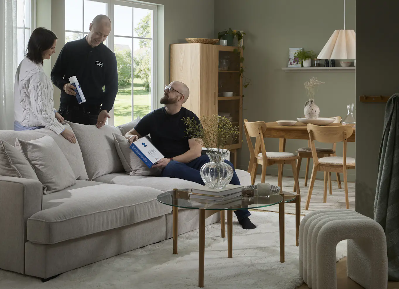 Servicepersonal från Mio med svarta kläder står i vardagsrum och pratar om möbelvårdsprodukter med kvinna intill och en man som sitter i en soffa. 