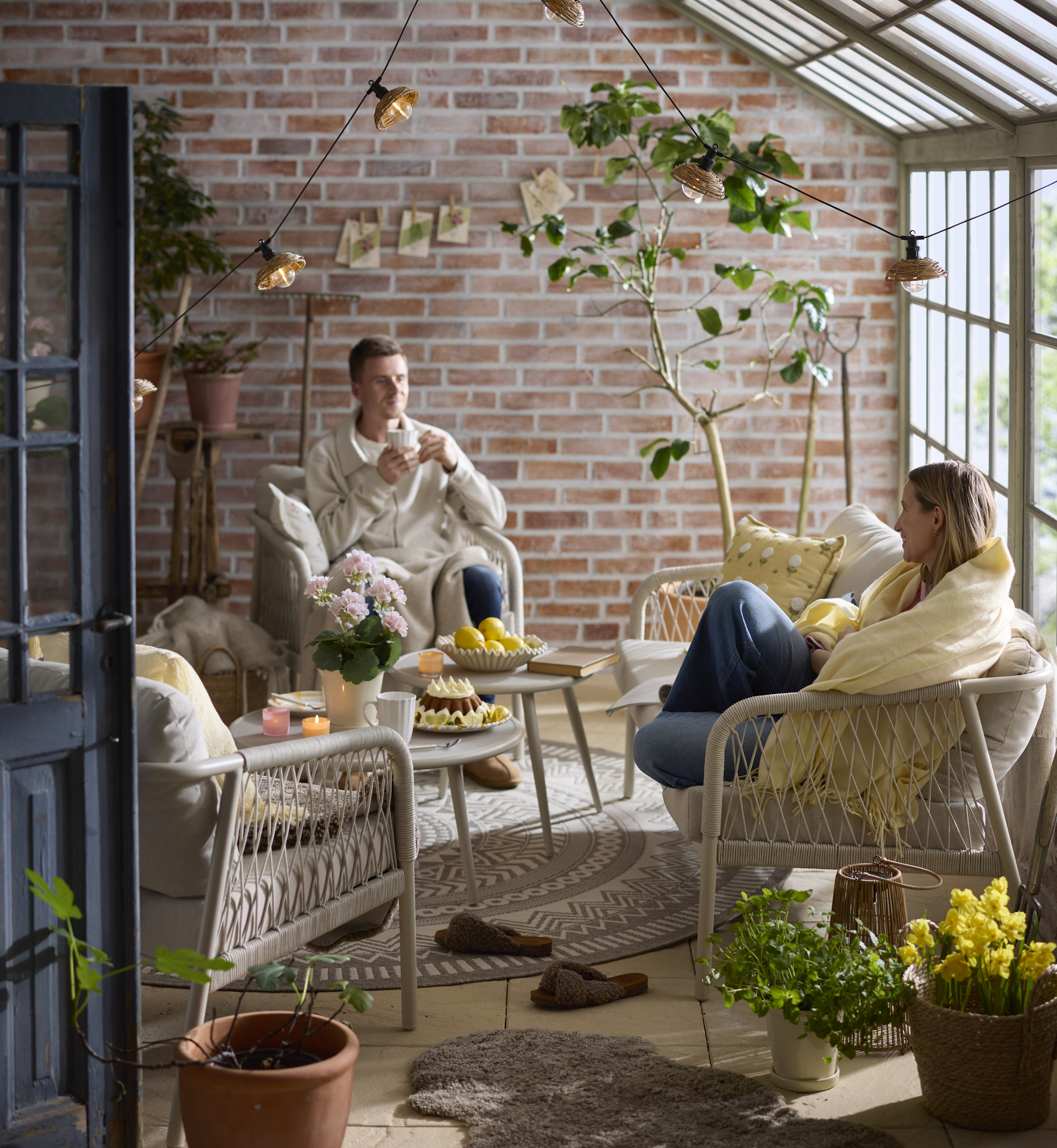 En man och en kvinna fikar i en beige loungergupp i ett orangeri fullt av växter och gula textilier.