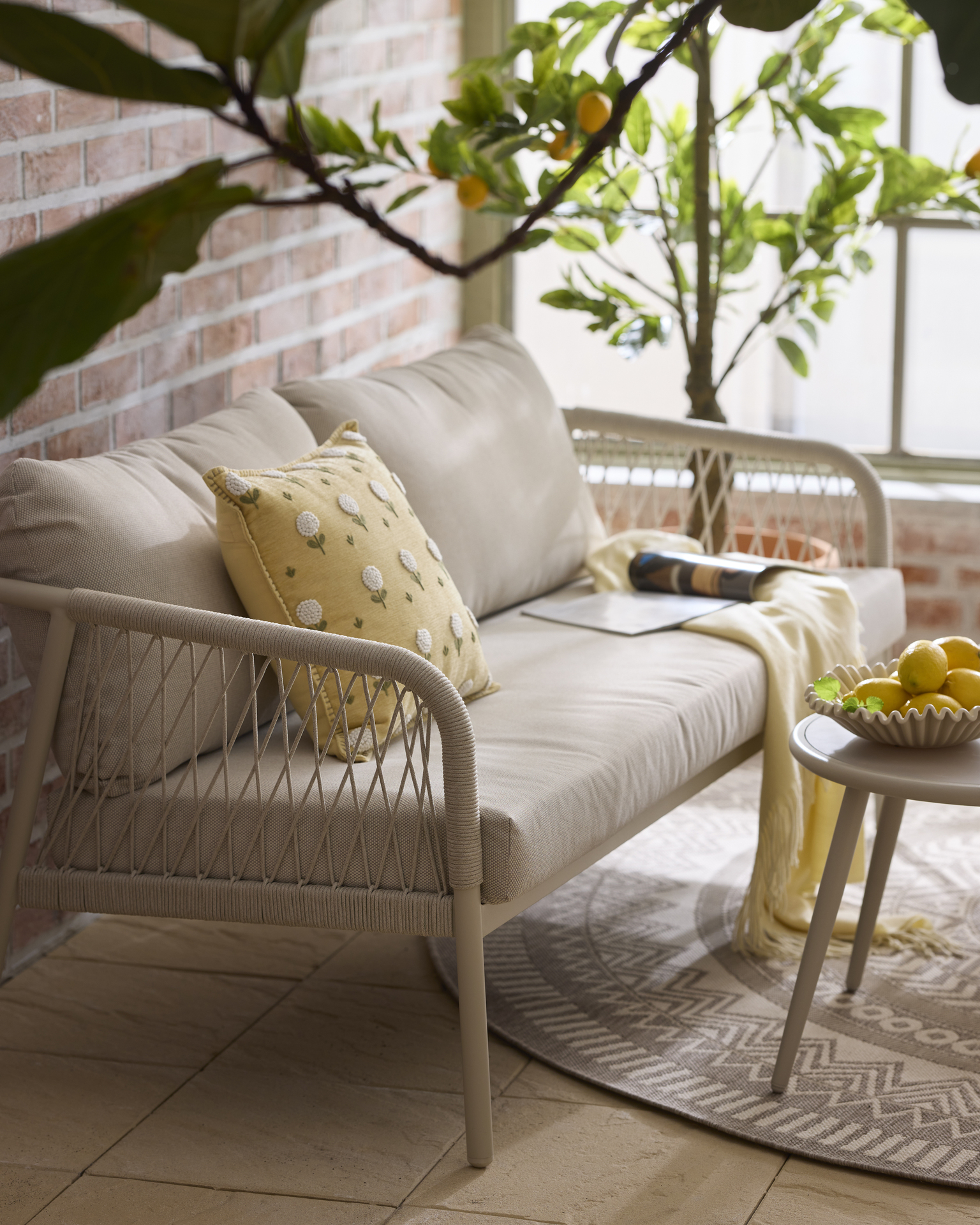 Detaljbild på en utesoffa i ett orangeri. I soffan ligger en kul kudde med blommönster och en gul pläd. I bakgrunden syns ett citronträd.