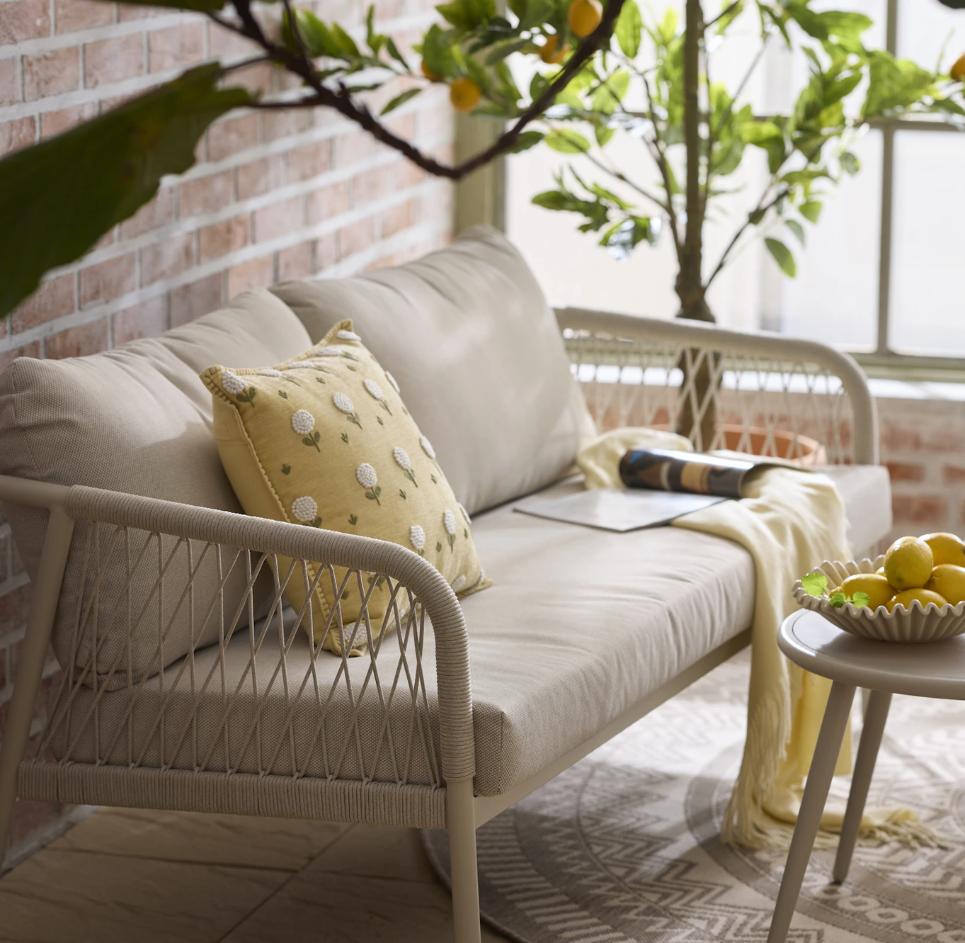 Detaljbild på en utesoffa i ett orangeri. I soffan ligger en kul kudde med blommönster och en gul pläd. I bakgrunden syns ett citronträd.