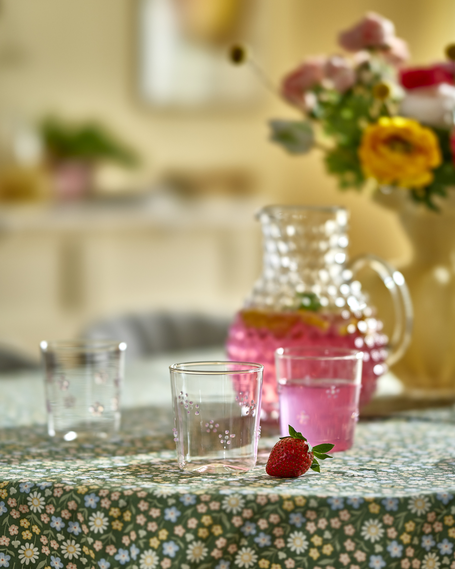 Dricksglas med blommor på står på en grön duk tillsammans med en karaff med rosa dryck i