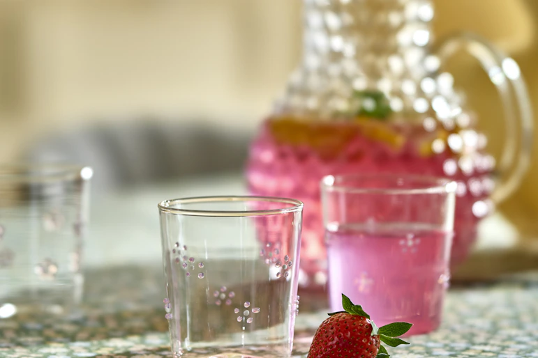 Dricksglas med blommor på står på en grön duk tillsammans med en karaff med rosa dryck i