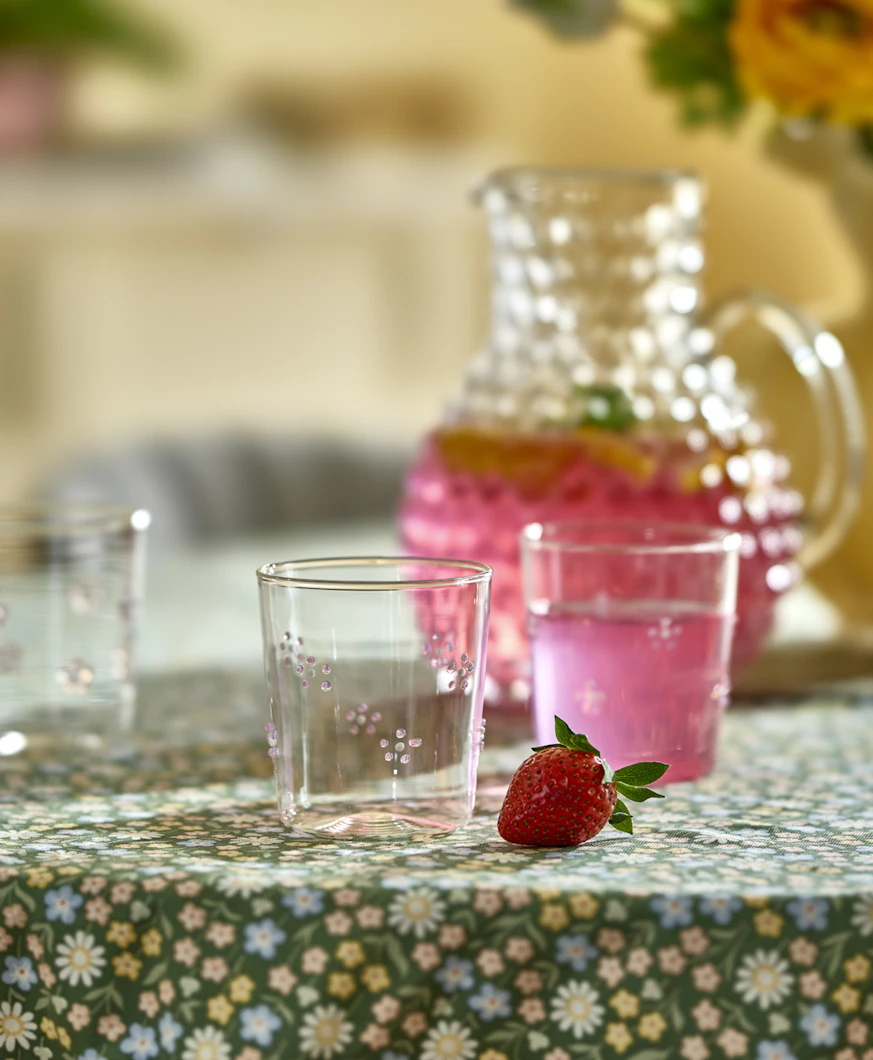 Dricksglas med blommor på står på en grön duk tillsammans med en karaff med rosa dryck i