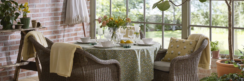 En utesoffgrupp med runt bord och fyra fåtöljer i konstrotting står i ett orangeri fyllt av växter.