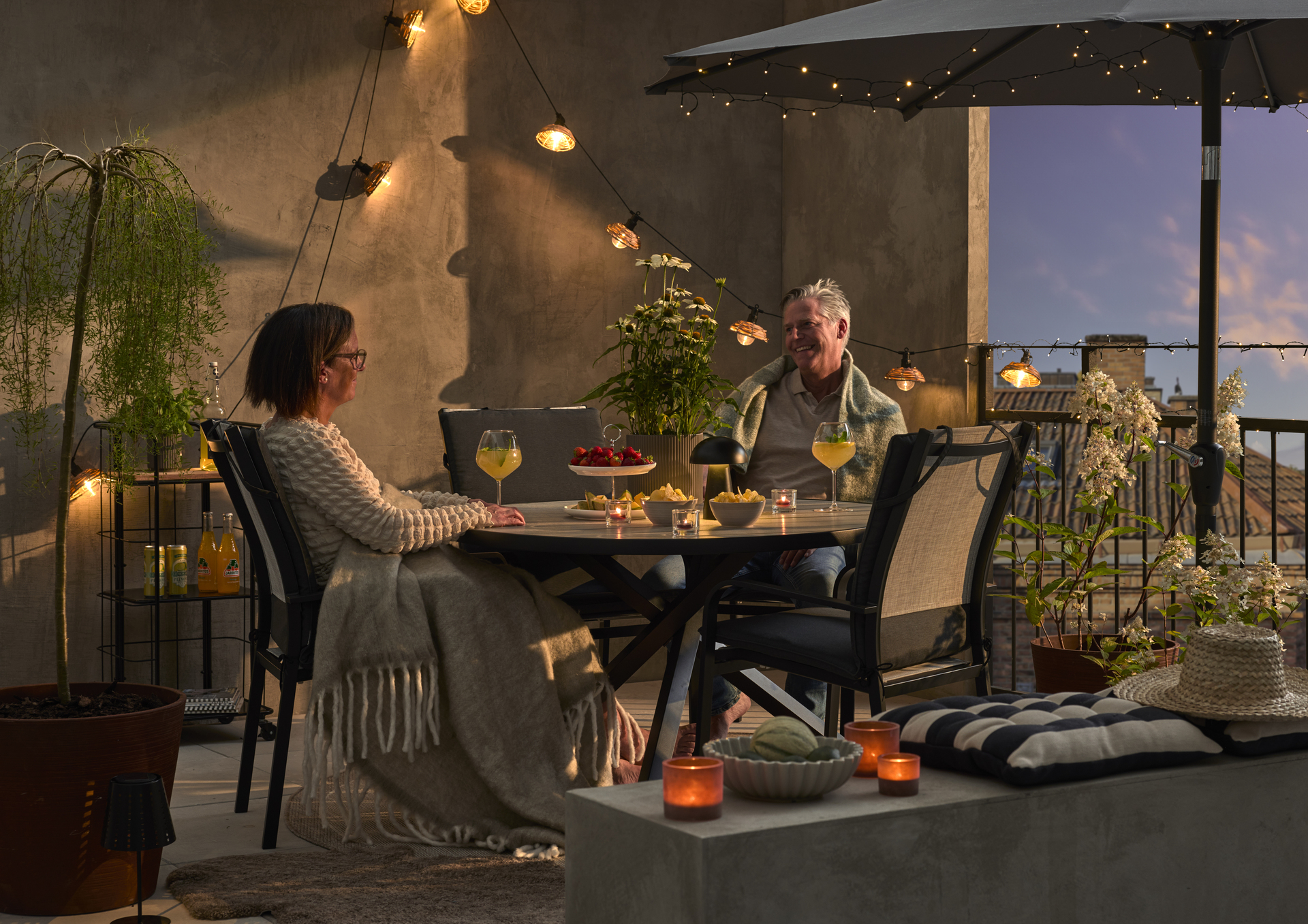I skymningsljus umgås en man och en kvinna i en matgrupp på en balkong. Balkongen är dekorerad med växter och solcellsbelysning.