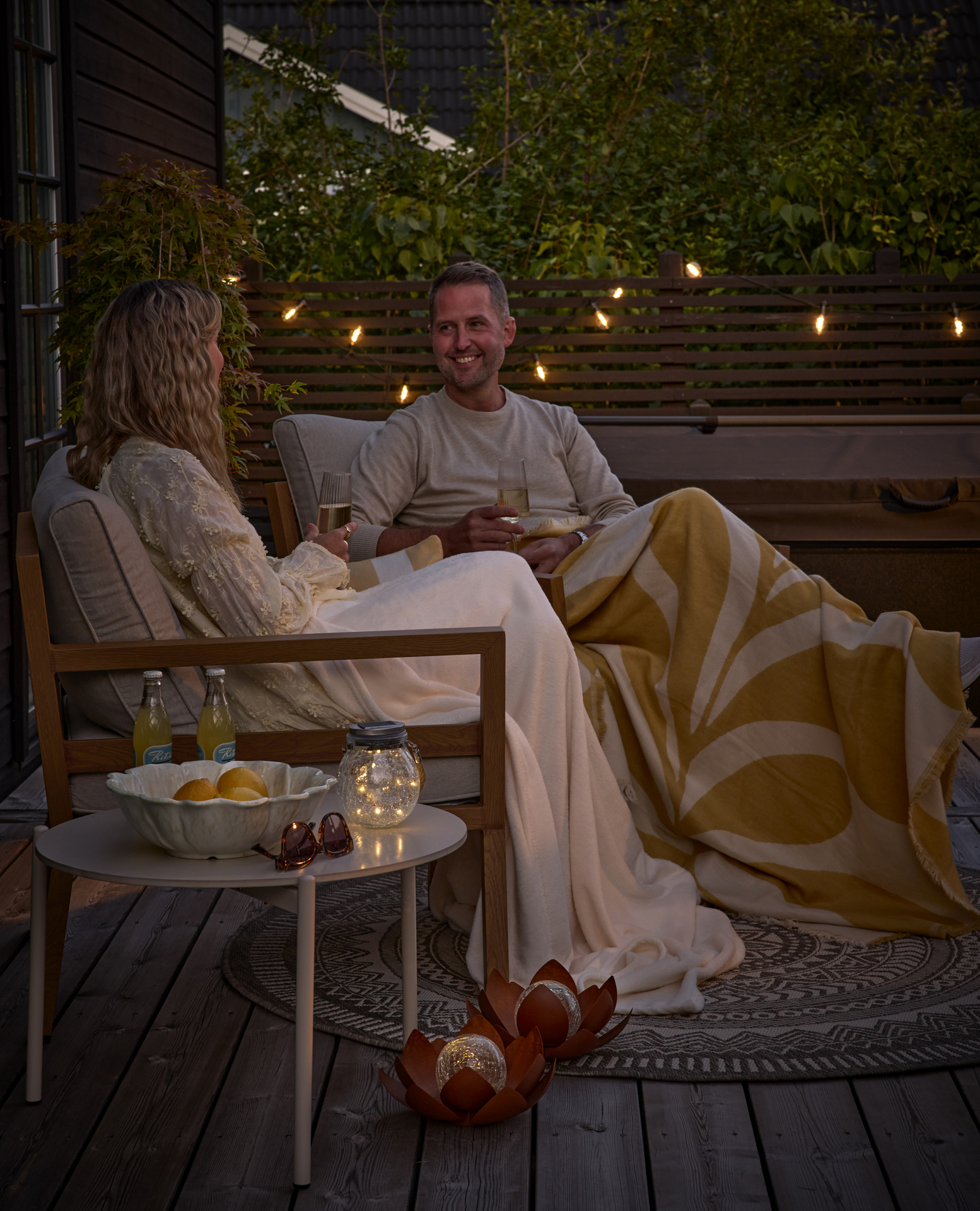 I skymningsljus sitter en man och en kvinna i utefåtöljer under varsin pläd och umgås.