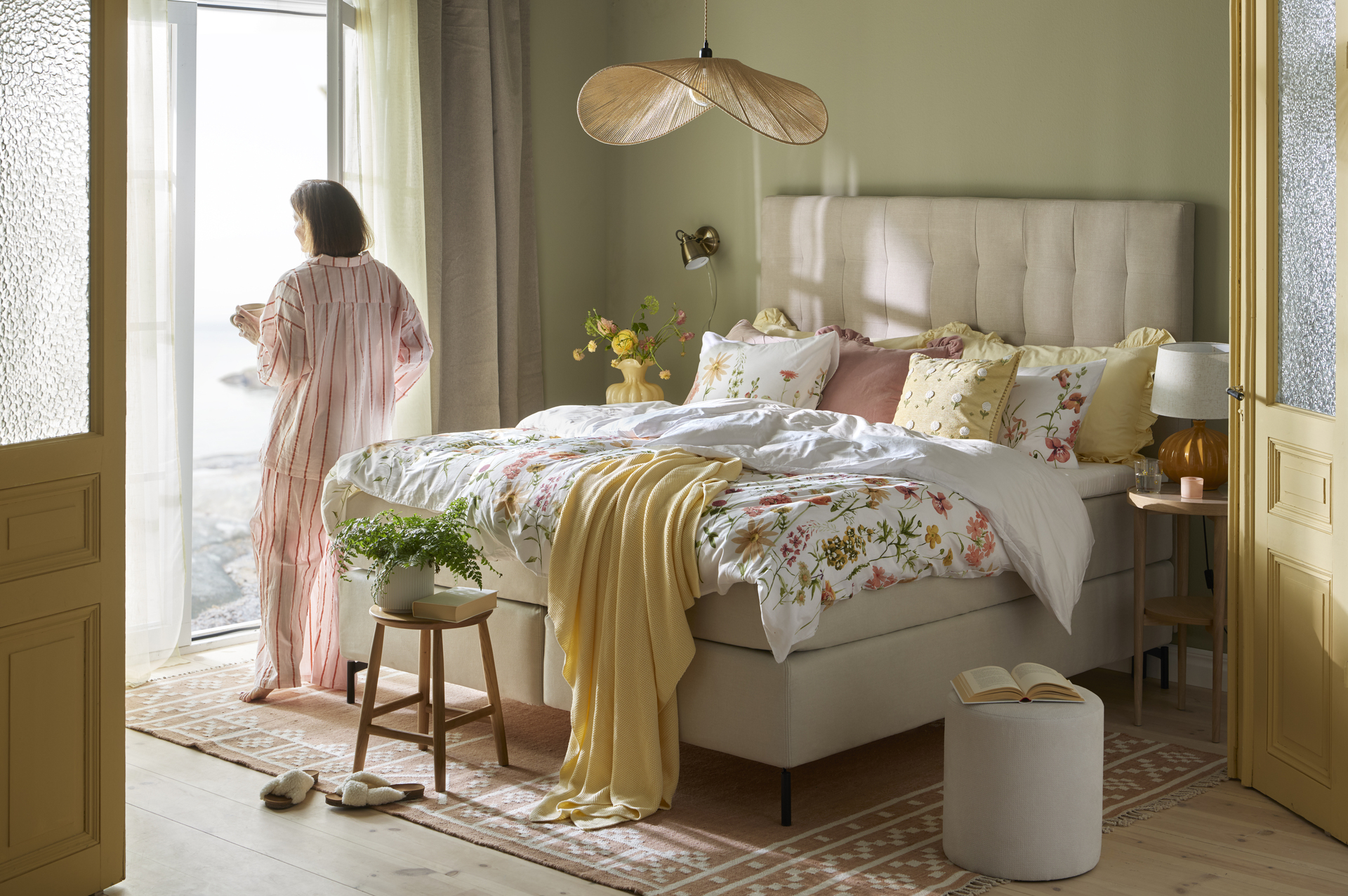 I ett salviagrönt sovrum står en beige säng med matchande sänggavel. Sängen är bäddad med påslakan i somriga färger och mönster och bredvid sängen står en kvinna och tittar ut genom fönstret.