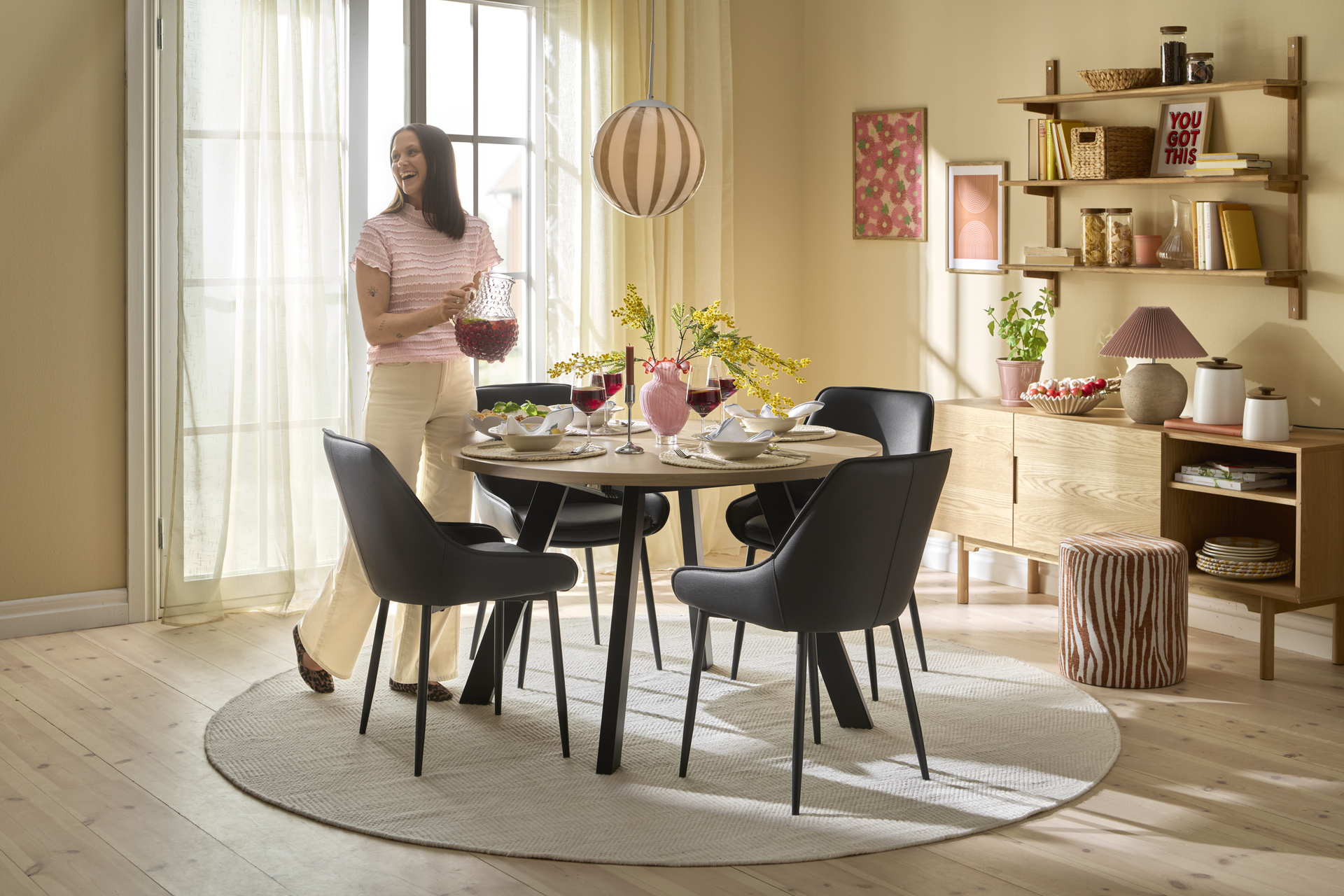 I ett rum målat i gult står ett runt matbord med fyra svarta stolar kring sig. Vid bordet står en kvinna och ler med en karaff i handen