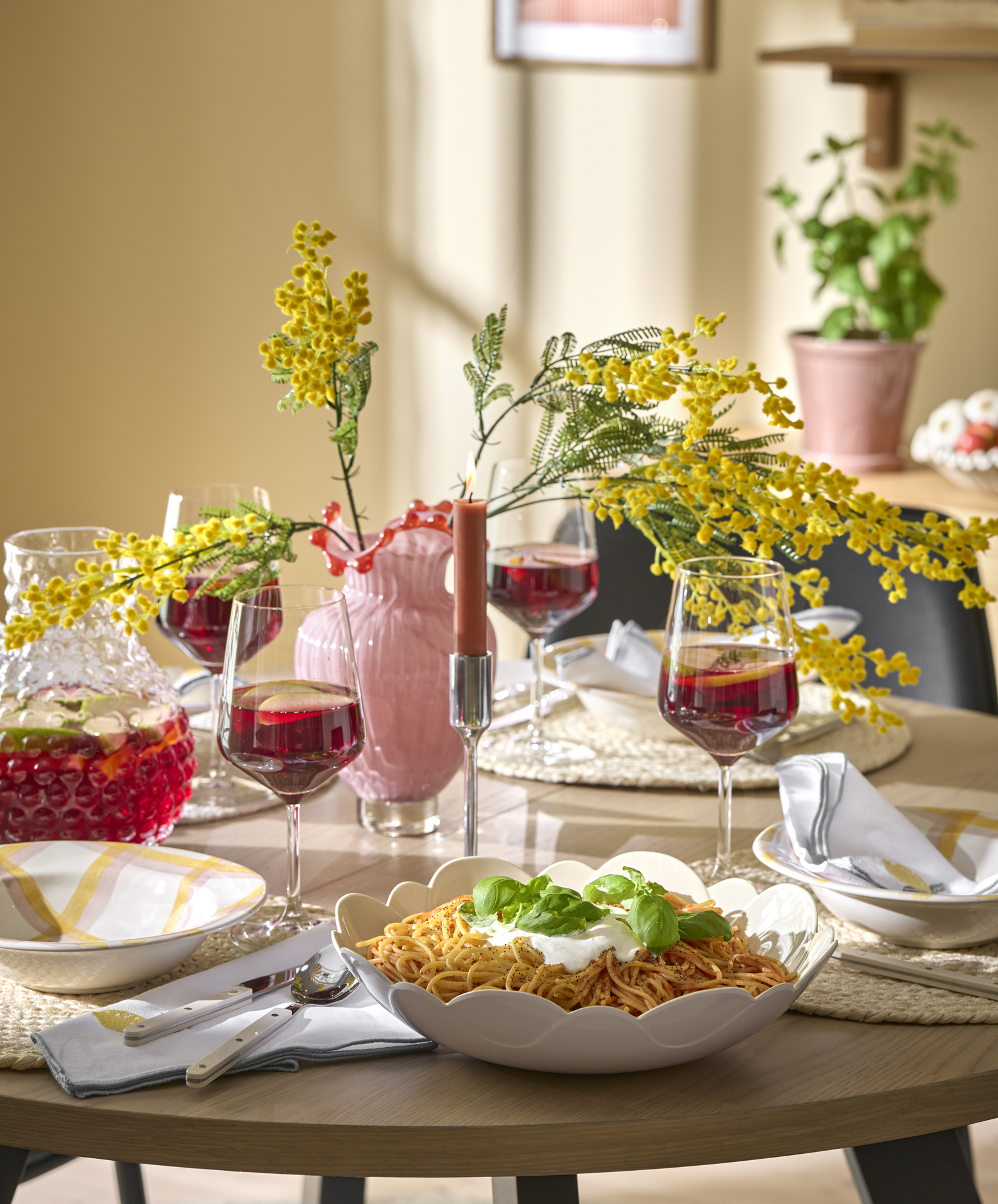 ett runt dukat bord med en skål pasta, vinglas och en rosa vas med gula blommor i