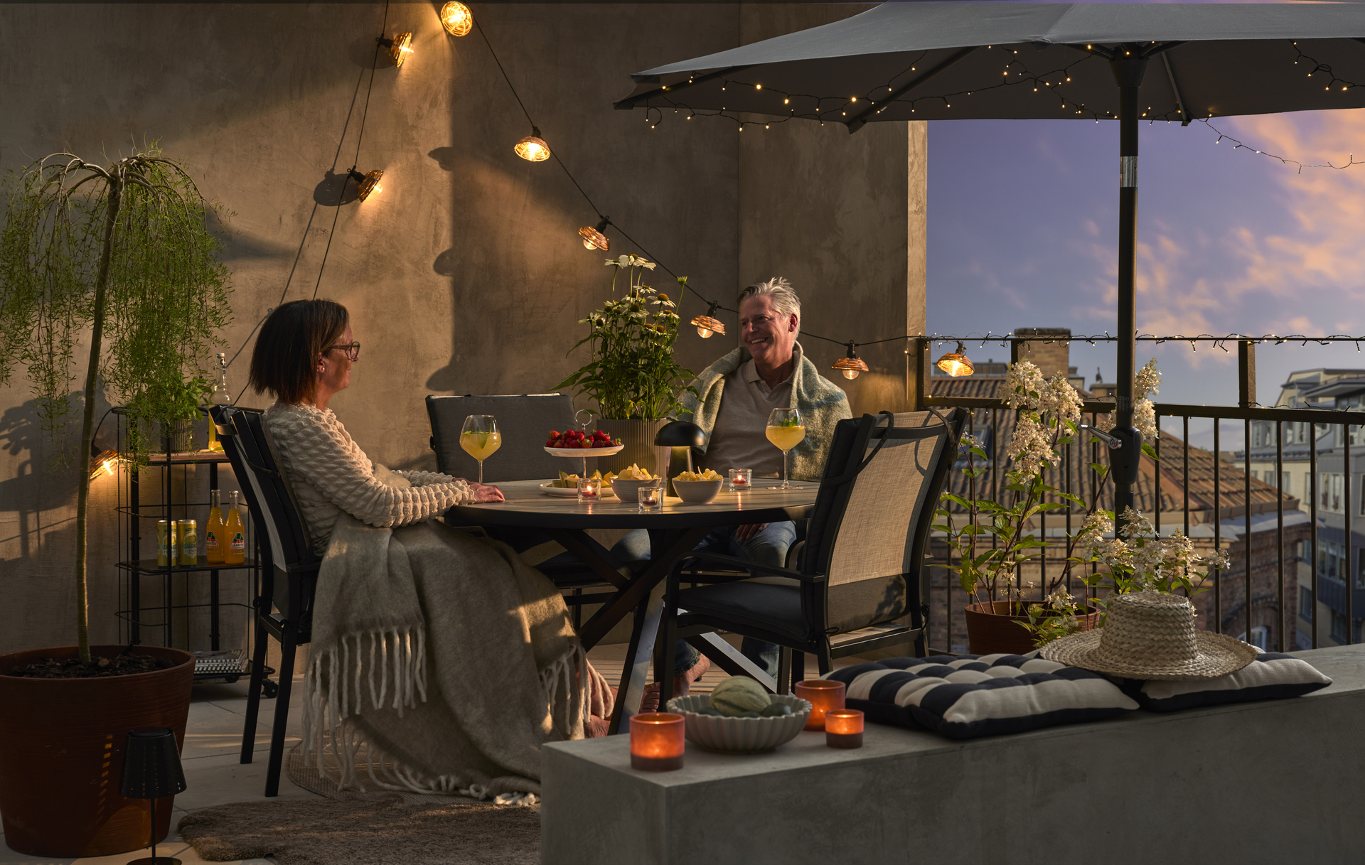 På en balkong upplyst med ljusslingor sitter en man och ler i skymningen