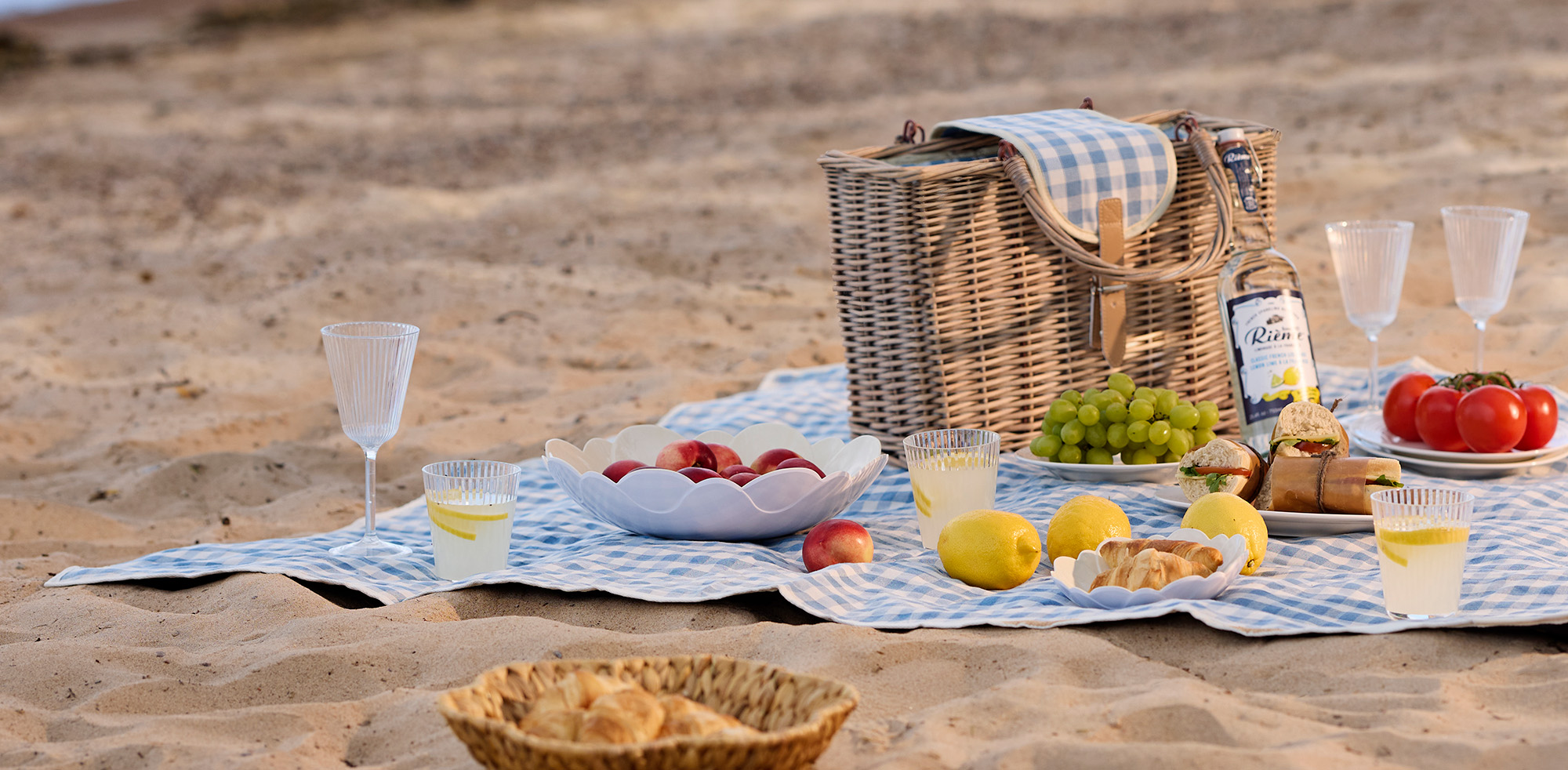 En blå picknickfilt ligger på en strand med en stor picknickkorg, glas, skålar och mat