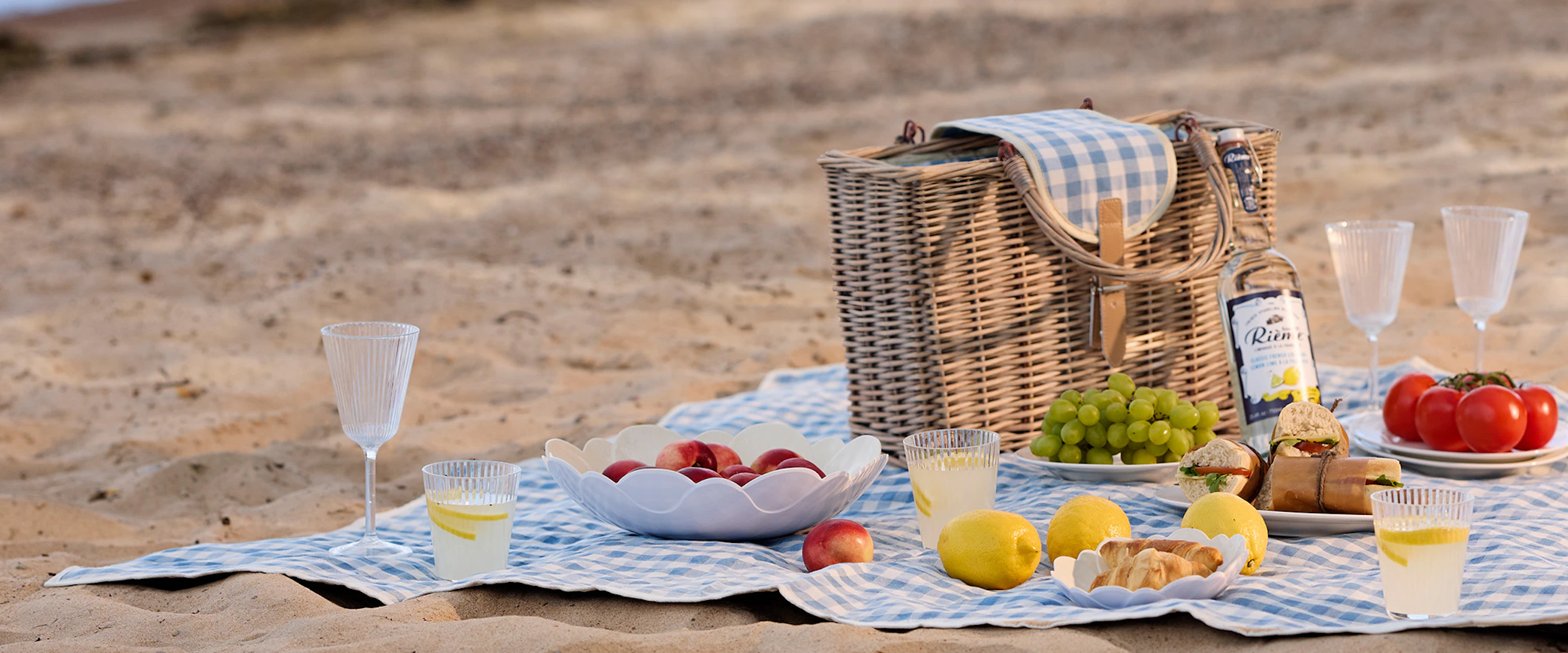 En blå picknickfilt ligger på en strand med en stor picknickkorg, glas, skålar och mat