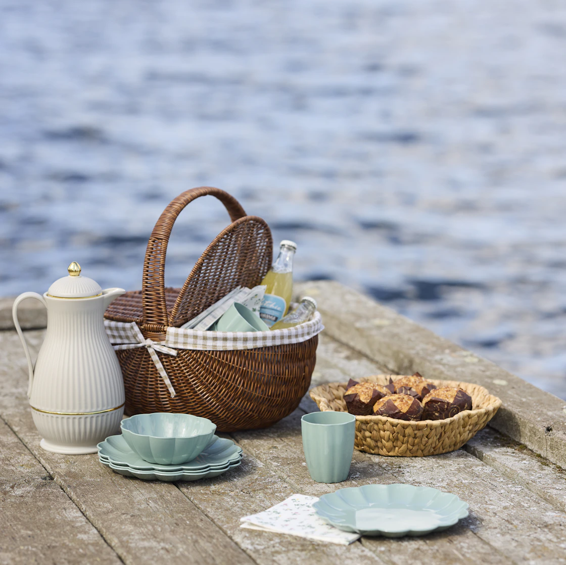 En picknickkorg med rutigt tyg inuti står på en brygga vid vattnet bredvid en termos, blå tallrikar och en korg med fika