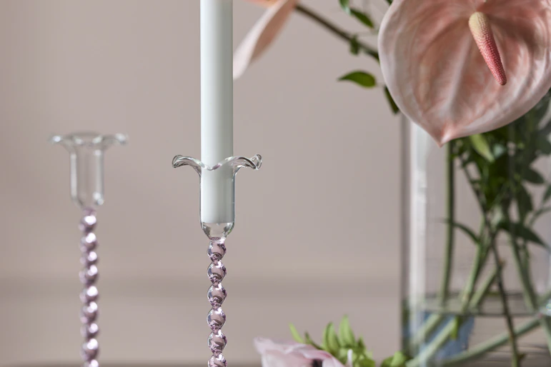 ett bord i mörkt trä står en ljusstake i rosa glas framför en vas med rosa blommor