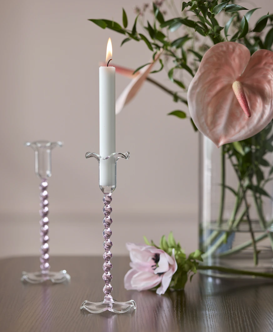 ett bord i mörkt trä står en ljusstake i rosa glas framför en vas med rosa blommor