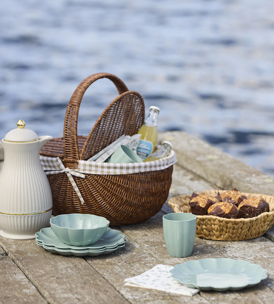 på en brygga vid vattnet står en picknickkorg samt blåa muggar och tallrikar, en korg med fika och en vit termos
