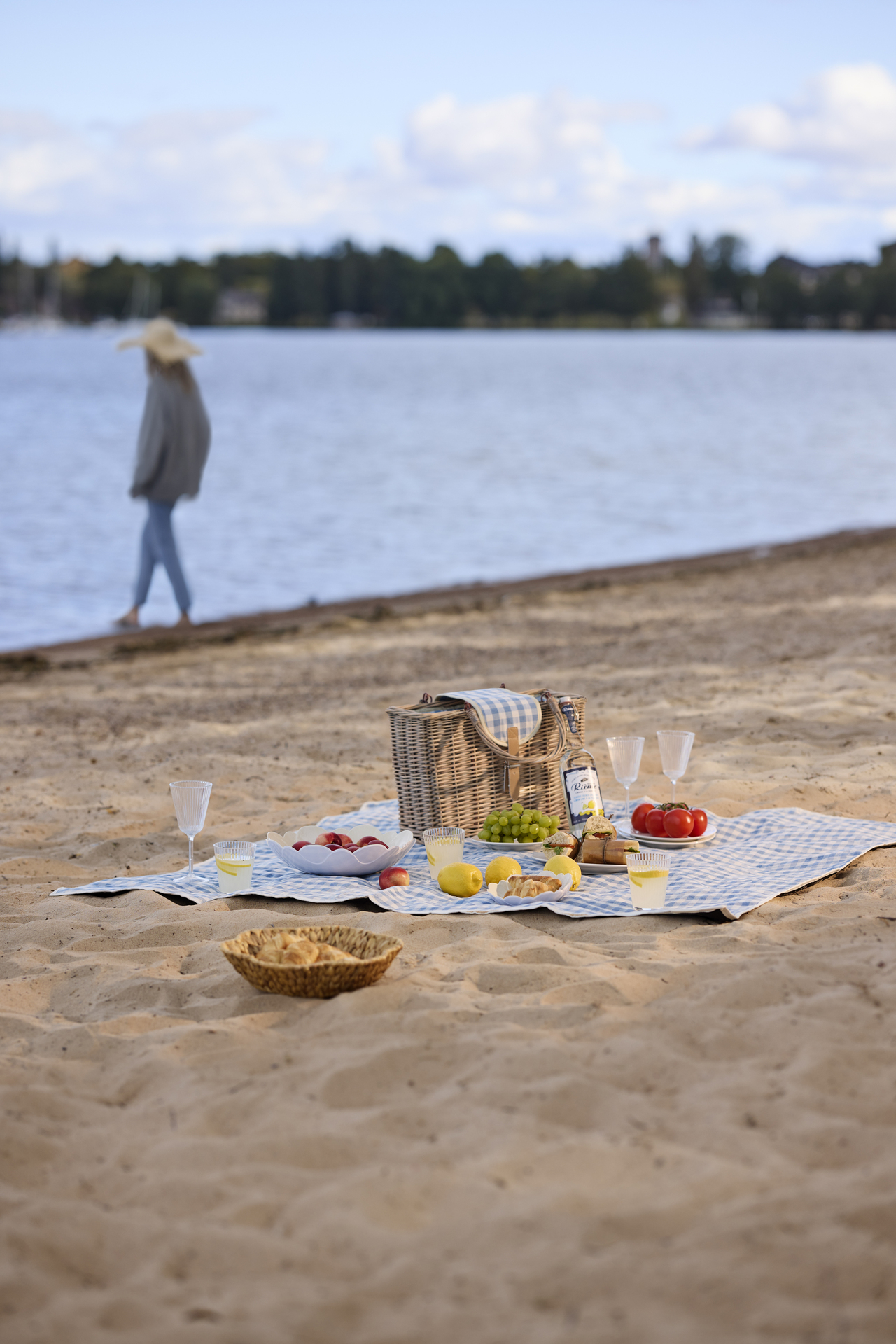 En blå picknickfilt ligger på en strand med en stor picknickkorg, glas, skålar och mat