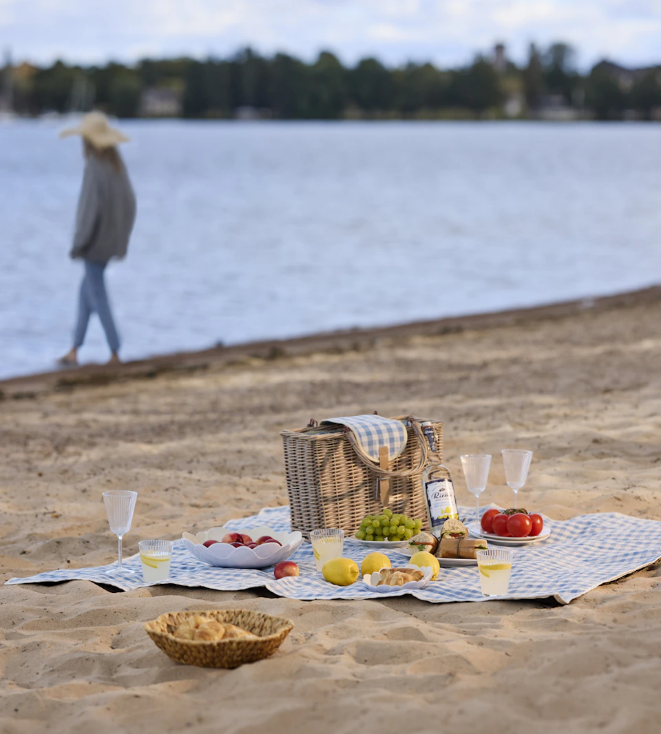 En blå picknickfilt ligger på en strand med en stor picknickkorg, glas, skålar och mat