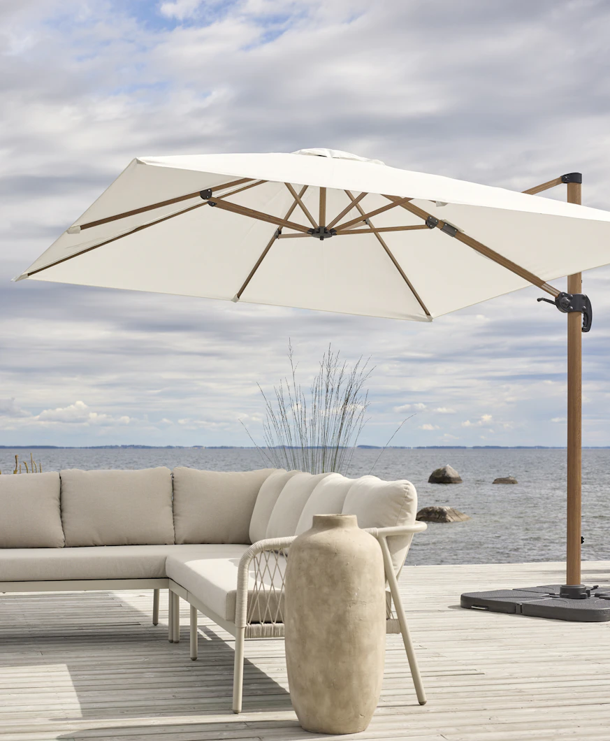På en altan intill vatten står ett stort vitt parasoll och en beige utesoffa.