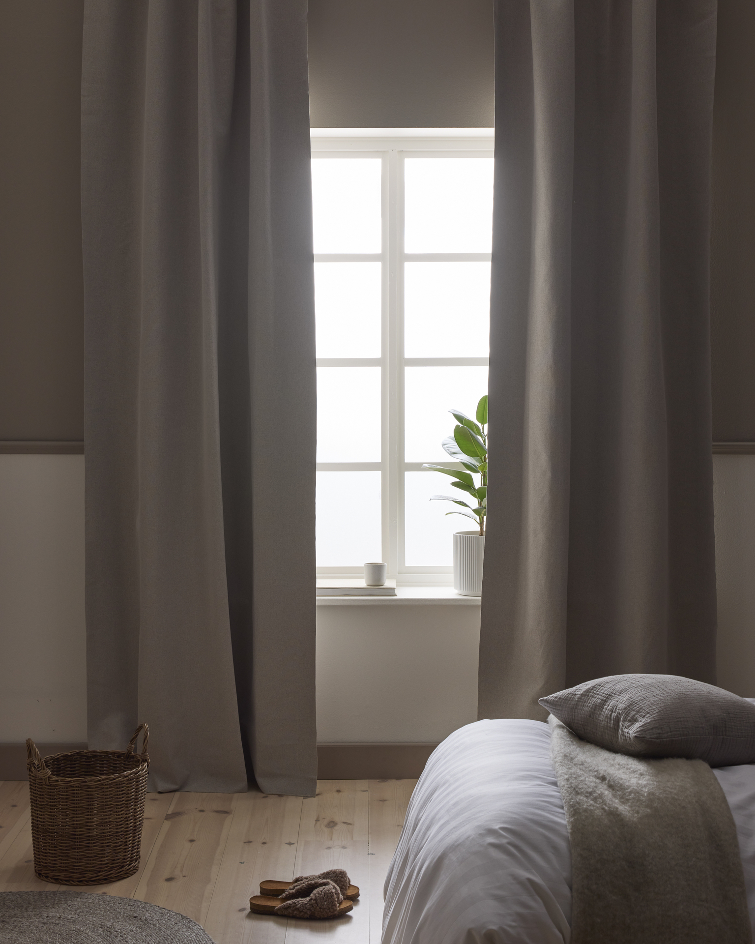 I ett sovrum i neutrala toner hänger beige mörkläggningsgardiner framför ett fönster