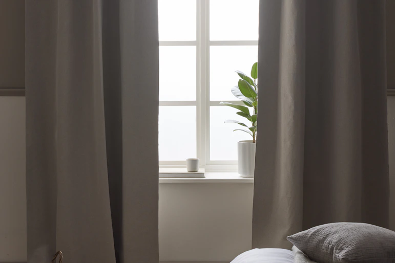 I ett sovrum i neutrala toner hänger beige mörkläggningsgardiner framför ett fönster