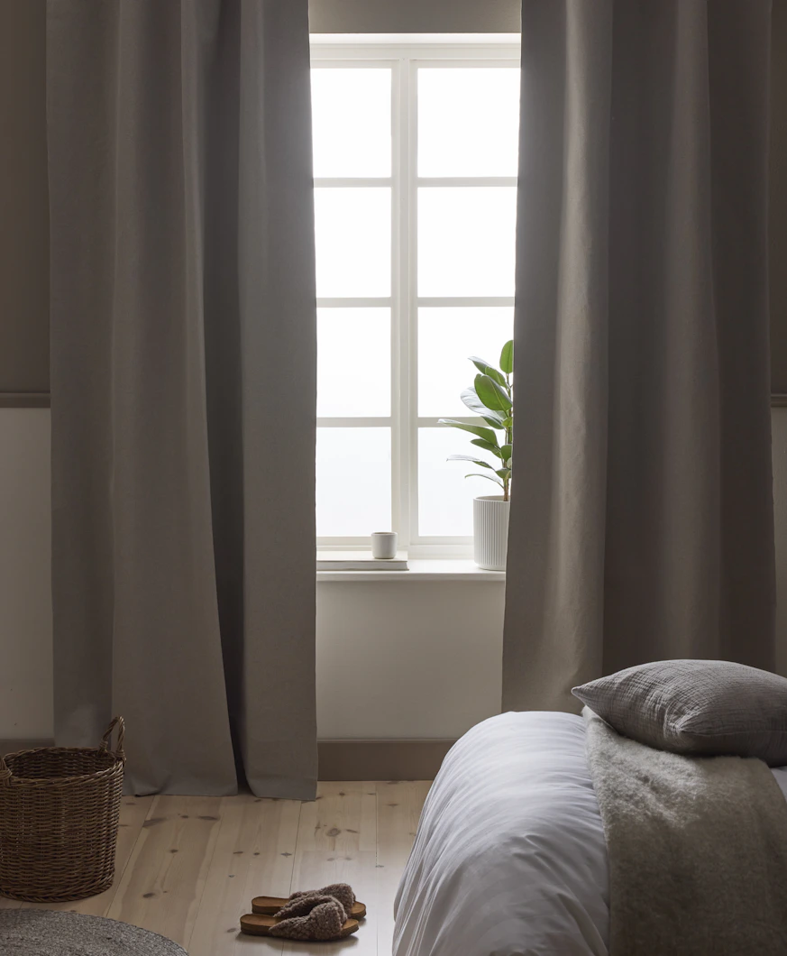 I ett sovrum i neutrala toner hänger beige mörkläggningsgardiner framför ett fönster