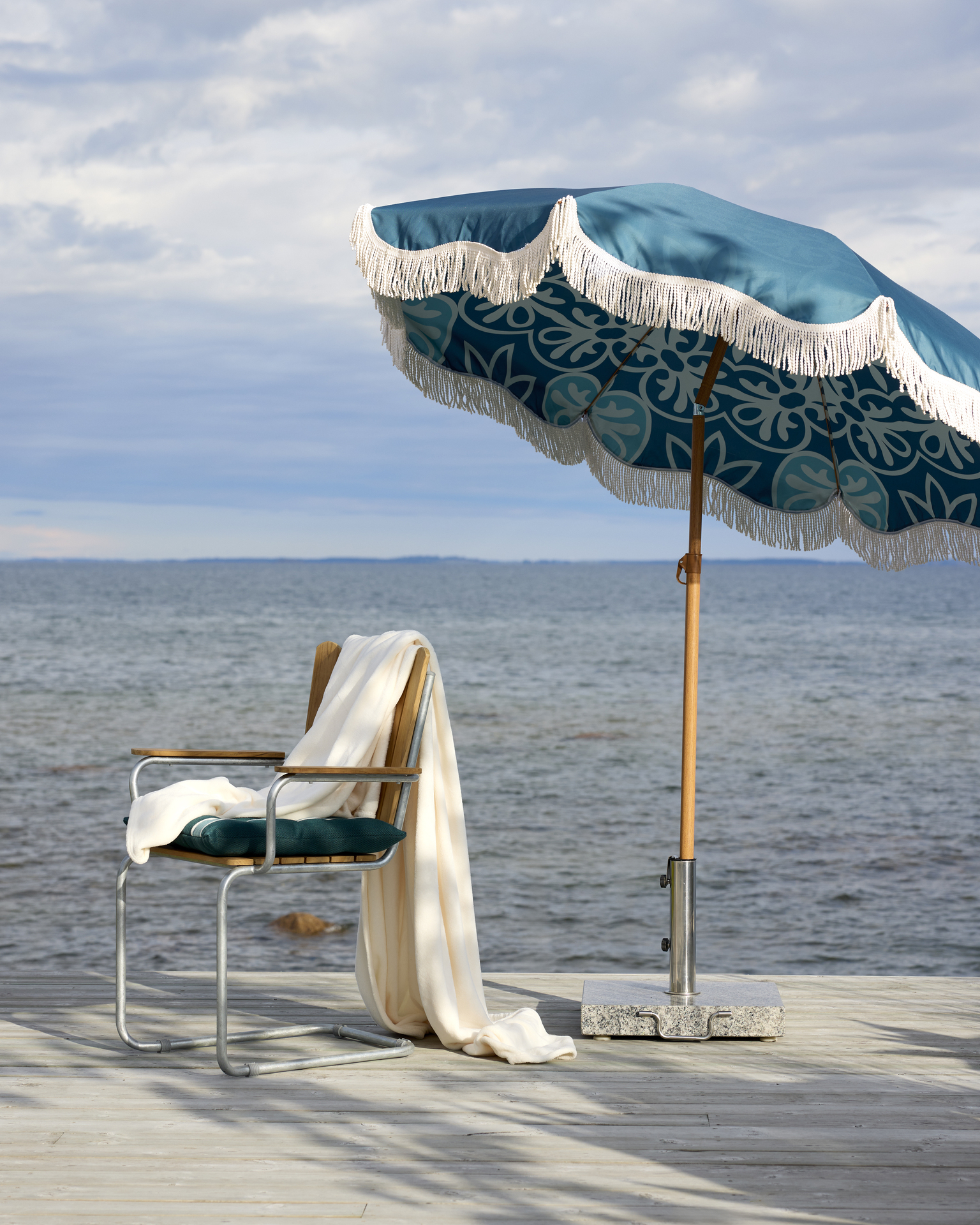 På en brygga står en stol under ett turkost parasoll med fransar