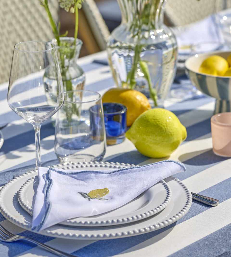 på ett bord med en randig duk i blått och vitt står vita tallrikar med linneservetter på samt vinglas