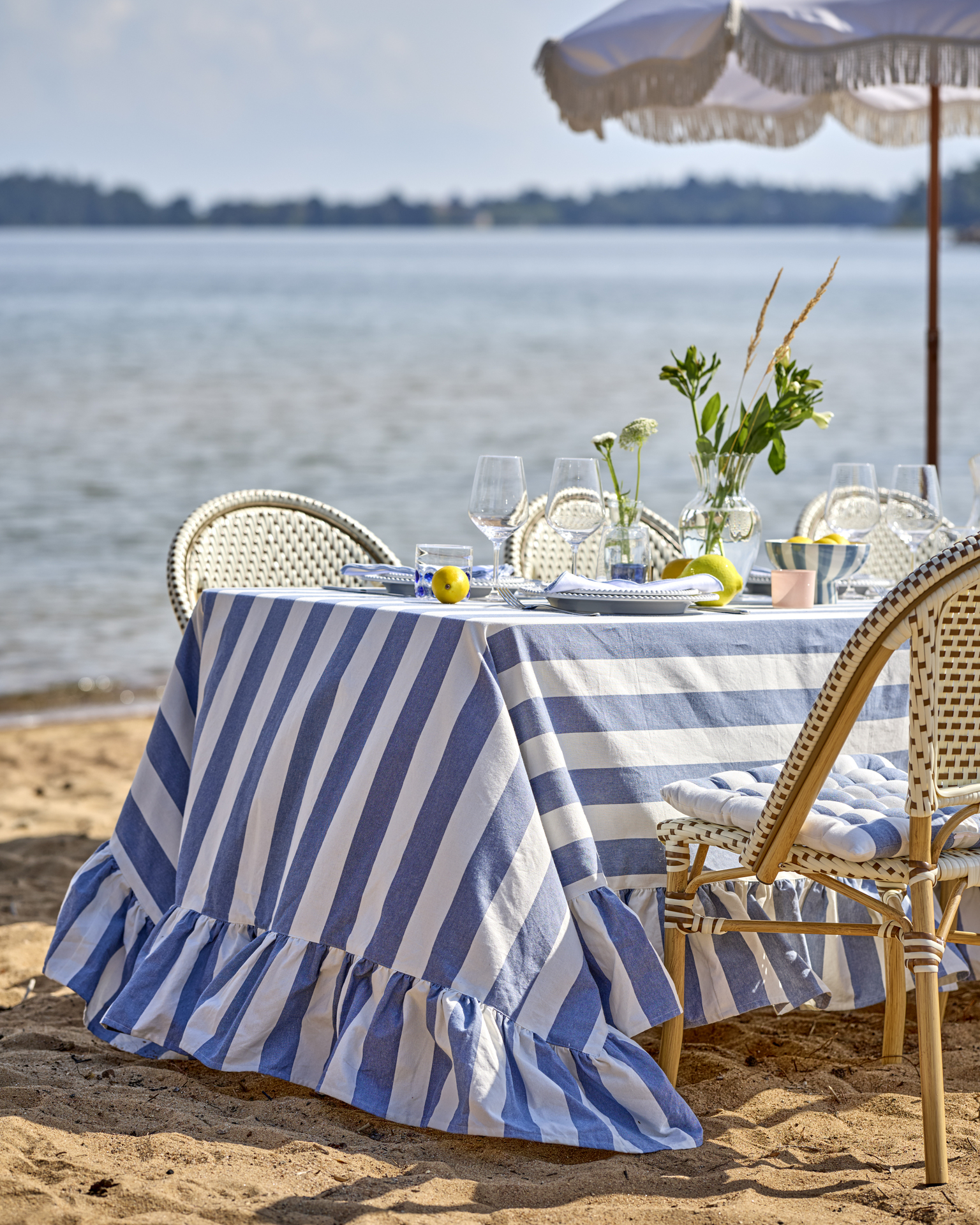 ett långt dukat bord med en blå- och vitrandig duk står på en strand vid vattnet