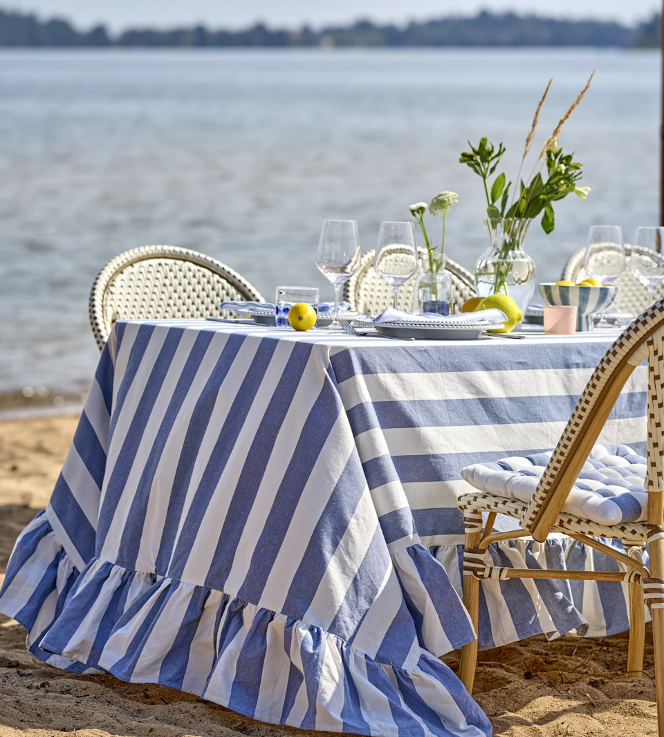 ett långt dukat bord med en blå- och vitrandig duk står på en strand vid vattnet