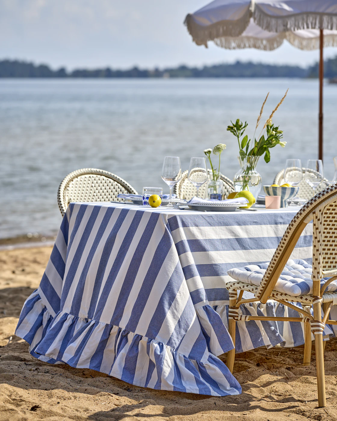 ett bord står dukat vid stranden med en randig blå duk, vitt porslin och vinglas