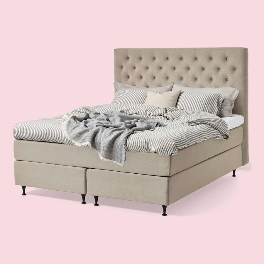 Beige kontinentalsäng med pikerad huvudgavel och beige bäddning mot en ljusrosa bakgrund. 