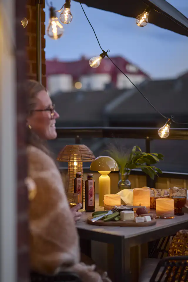 På en liten, mysig balkong sitter en kvinna i skymningen. Balkongen är pyntad med massor av lampor och ljuslyktor
