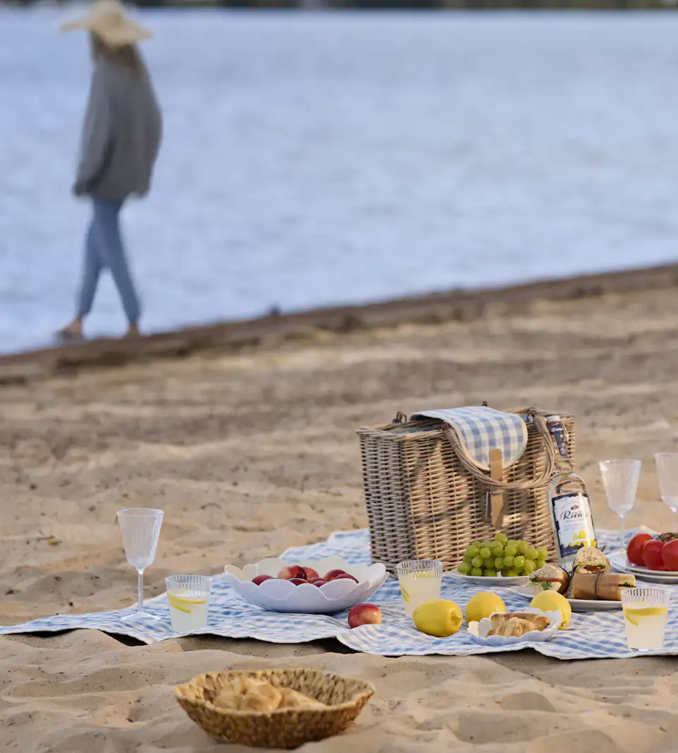 På en strand ligger en blåvit rutig picknickfilt. På filten är en festli picknick framdukad