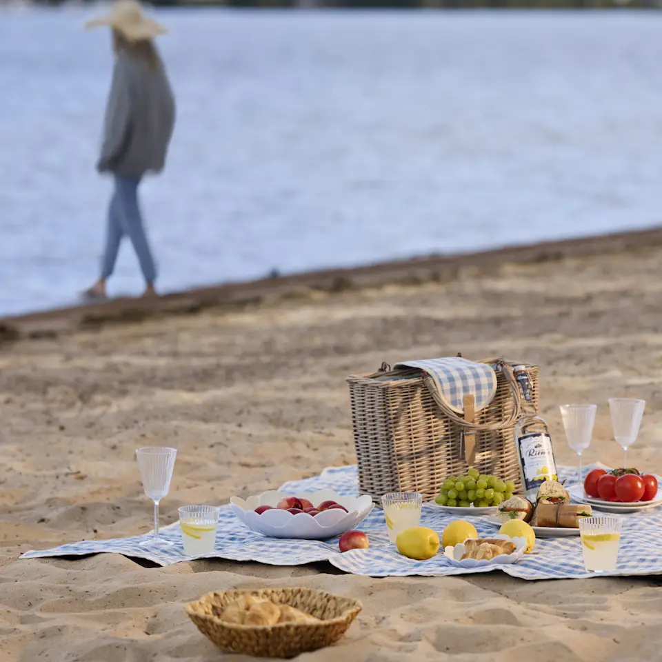 På en strand ligger en blåvit rutig picknickfilt. På filten är en festli picknick framdukad