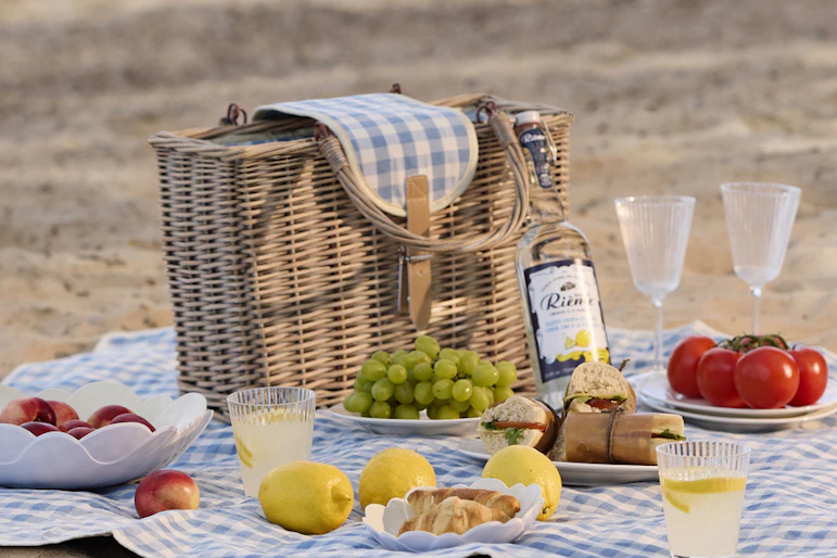På en strand ligger en blåvit rutig picknickfilt. På filten är en festli picknick framdukad