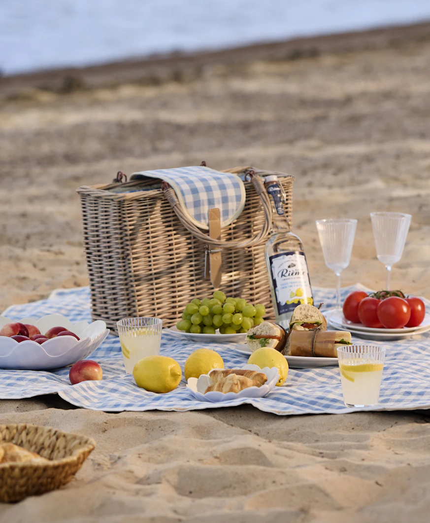 På en strand ligger en blåvit rutig picknickfilt. På filten är en festli picknick framdukad