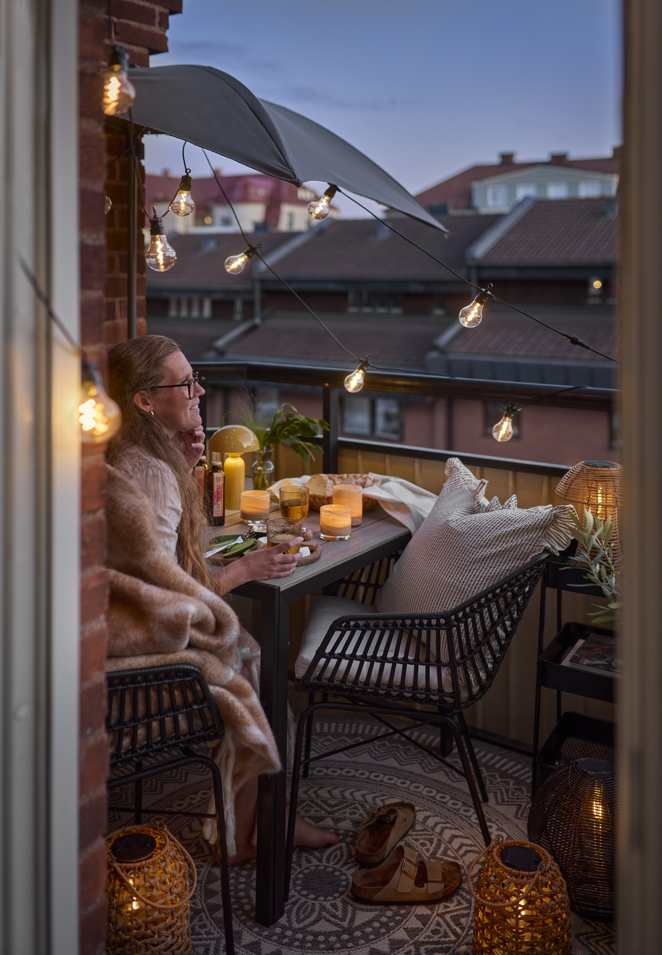 På en liten, mysig balkong sitter en kvinna i skymningen. Balkongen är pyntad med massor av lampor och ljuslyktor