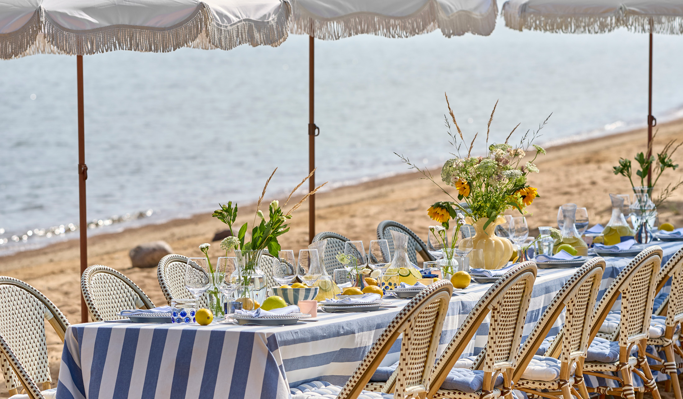 ett bord står dukat på en strand med en randig duk, porslin och glas i olika utföranden och en stor gul vas med blommor