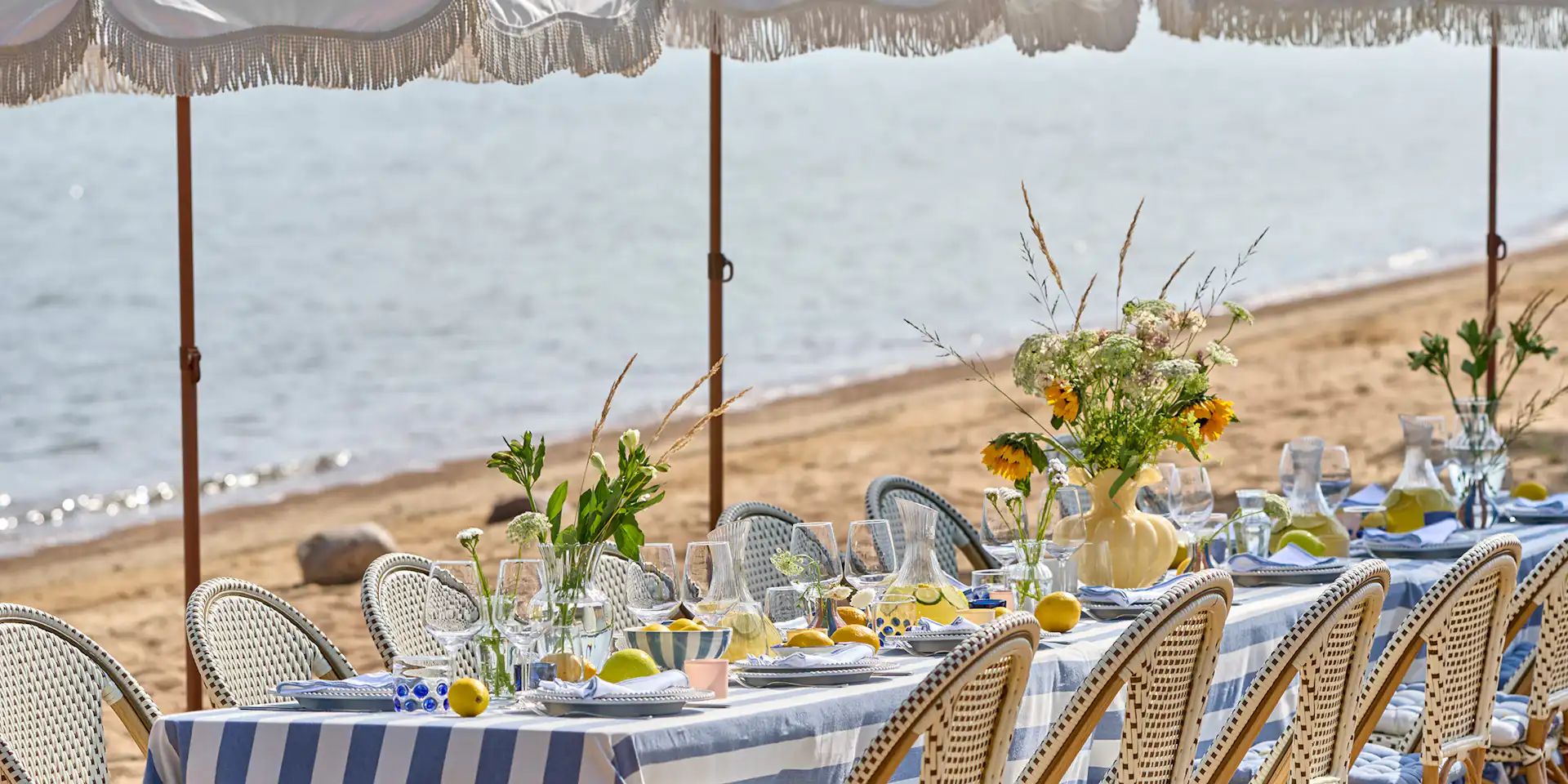 ett bord står dukat på en strand med en randig duk, porslin och glas i olika utföranden och en stor gul vas med blommor
