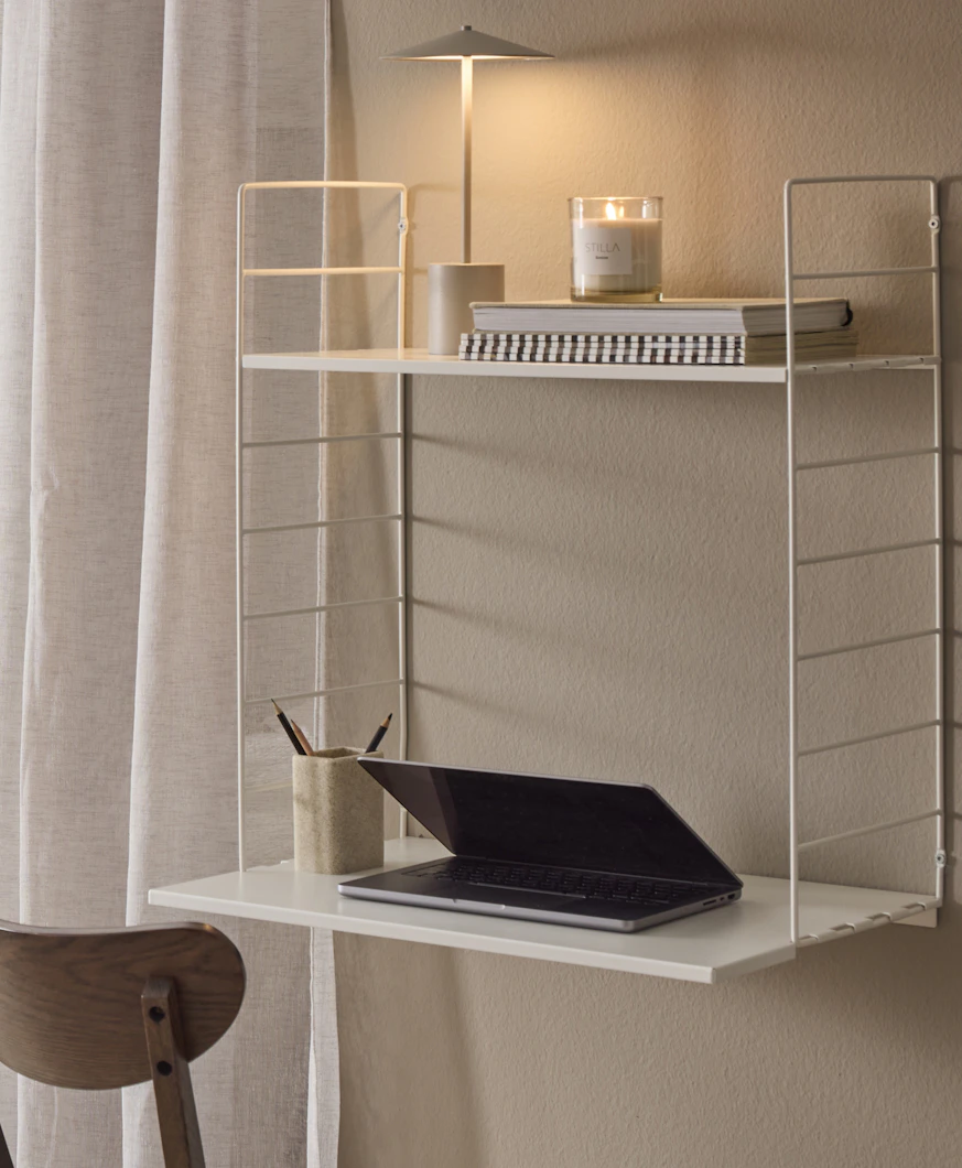 En laptop, en lampa, några böcker och ett doftljus står i en vit vägghylla som används som skrivbord.