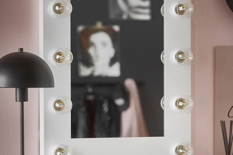 En vit sminkspegel med tolv ljuskällor runt ramen hänger på en rosa vägg.