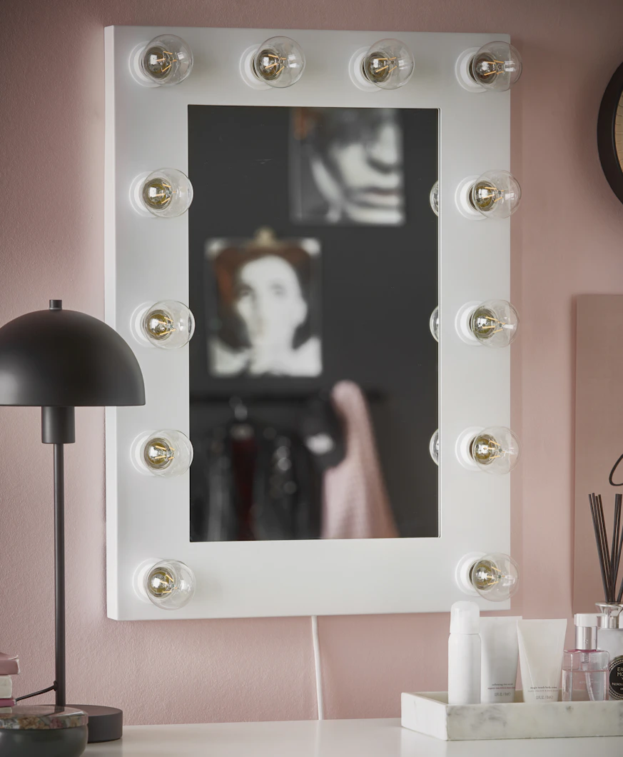 En vit sminkspegel med tolv ljuskällor runt ramen hänger på en rosa vägg.
