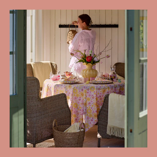En tjej i rosa skjorta står i ett uterum bakom ett bord som är dukat med en rosa och gul duk, porslin och påskdekorationer