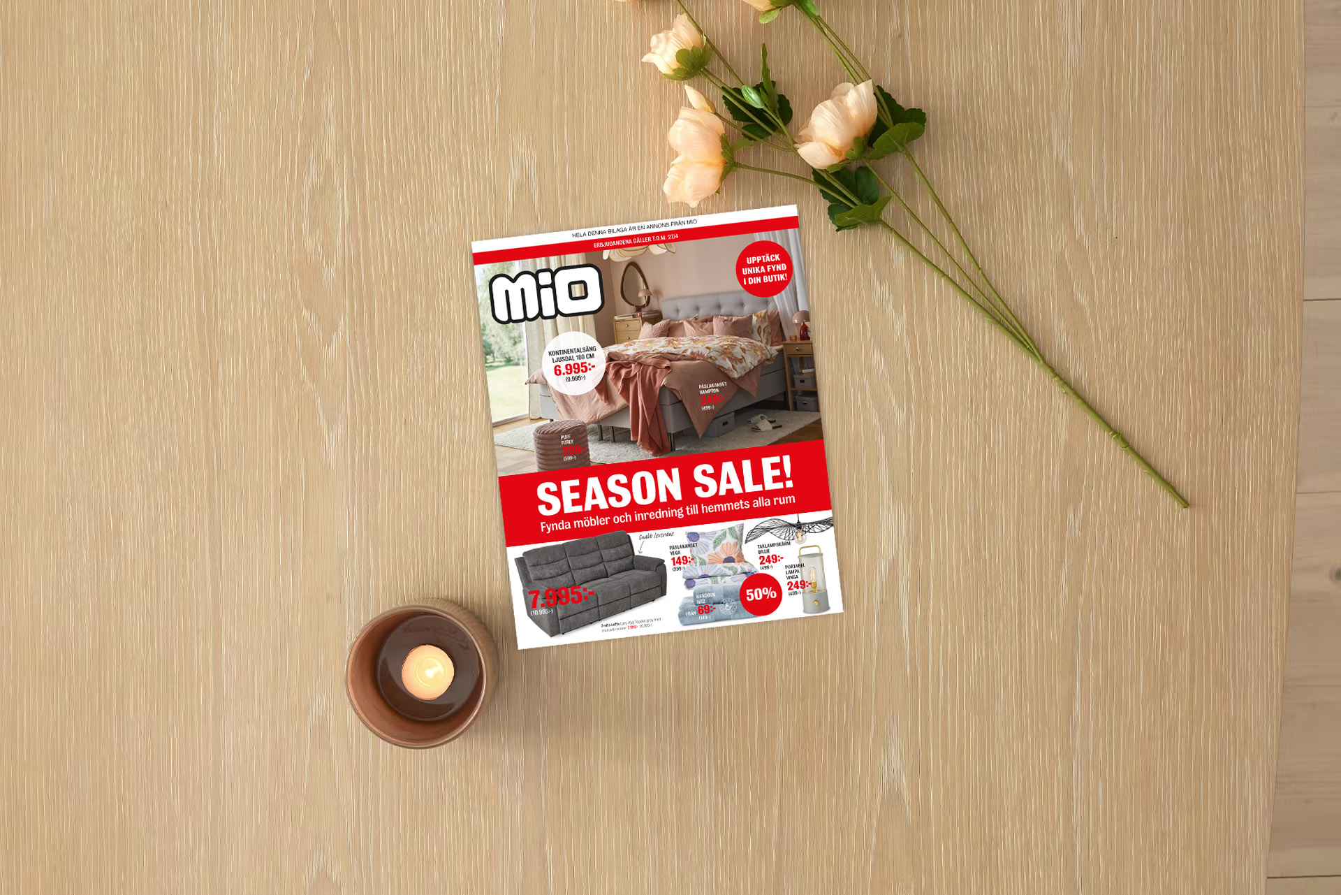 På ett ljust träbord ligger kampanjbladet för Season Sale intill ett ljus och en blomma.
