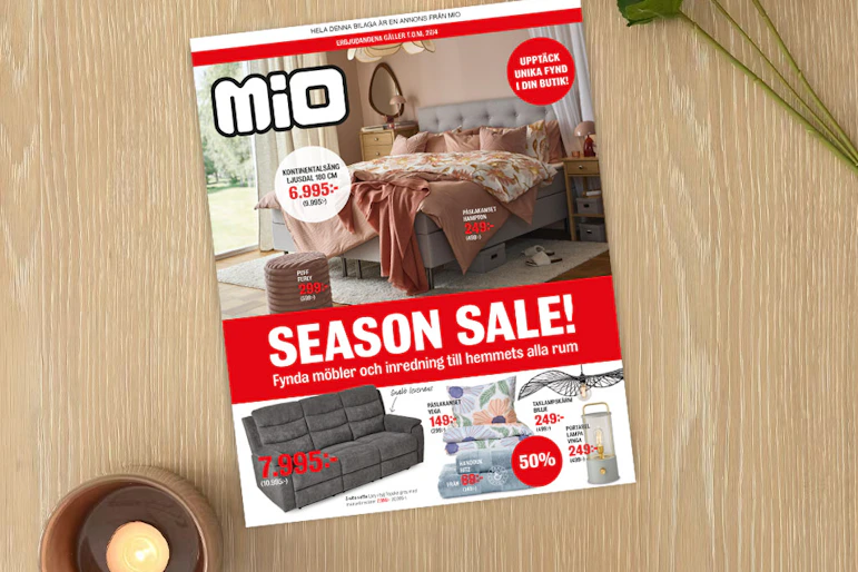 På ett ljust träbord ligger kampanjbladet för Season Sale intill ett ljus och en blomma.