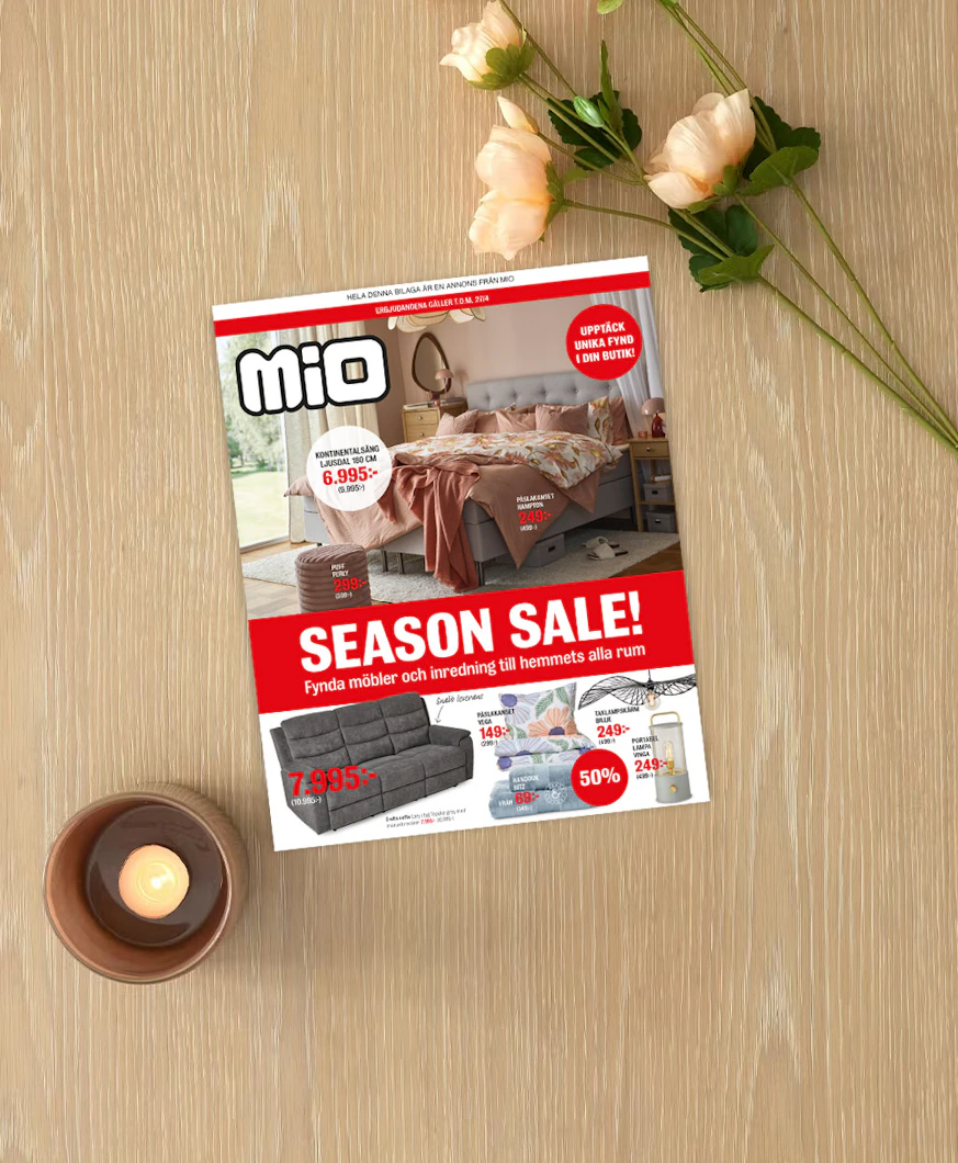 På ett ljust träbord ligger kampanjbladet för Season Sale intill ett ljus och en blomma.